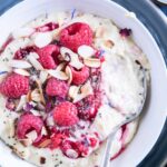 Vanillepudding-Porridge mit warmen Himbeeren in einer weißen Schüssel auf einem blauen Teller im Top View aus der Nähe mit einem Löffel, der im Haferbrei steckt und die Cremigkeit zeigt