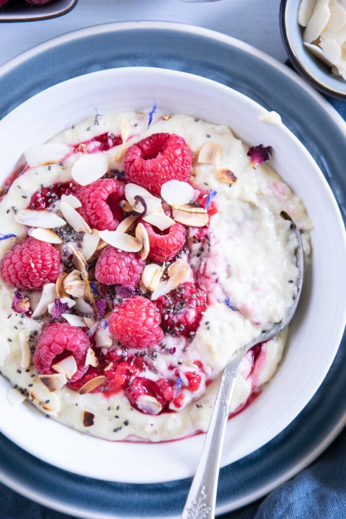 Vanillepudding-Porridge mit warmen Himbeeren in einer weißen Schüssel auf einem blauen Teller im Top View aus der Nähe mit einem Löffel, der im Haferbrei steckt und die Cremigkeit zeigt