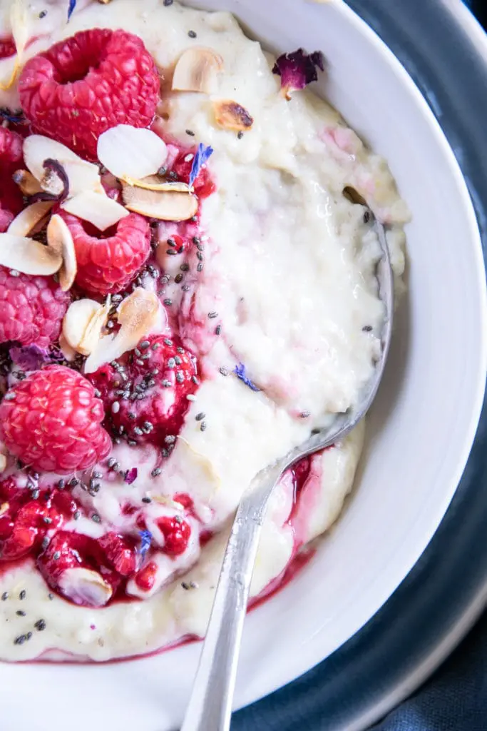 Vanillepudding-Porridge mit warmen Himbeeren in einer weißen Schüssel auf einem blauen Teller im Top View aus der Nähe mit einem Löffel, der im Haferbrei steckt und die Cremigkeit zeigt