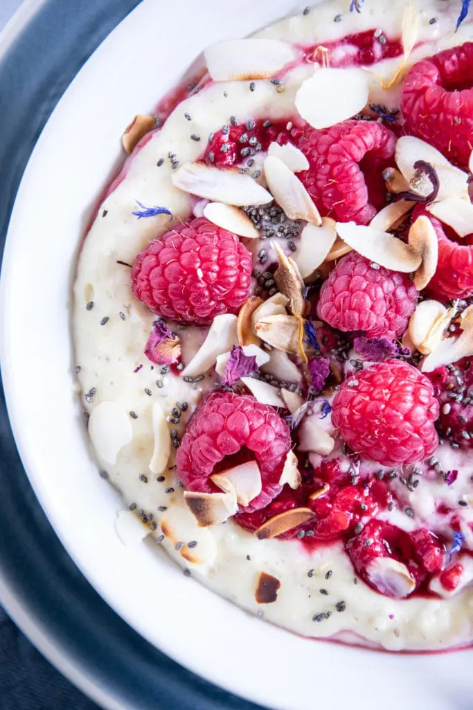 Vanillepudding-Porridge mit warmen Himbeeren in einer weißen Schüsse auf einem blauen Teller im Top View angeschnitten von rechts, mit Himbeeren, Mandelblättchen, Chiasamen und Blüten als Topping