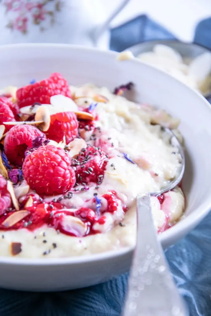 Vanillepudding-Porridge mit warmen Himbeeren in einer weißen Schüssel auf einem blauen Teller im 45-Grad-Winkel aus der Nähe mit einem Löffel, der im Haferbrei steckt und die Cremigkeit zeigt