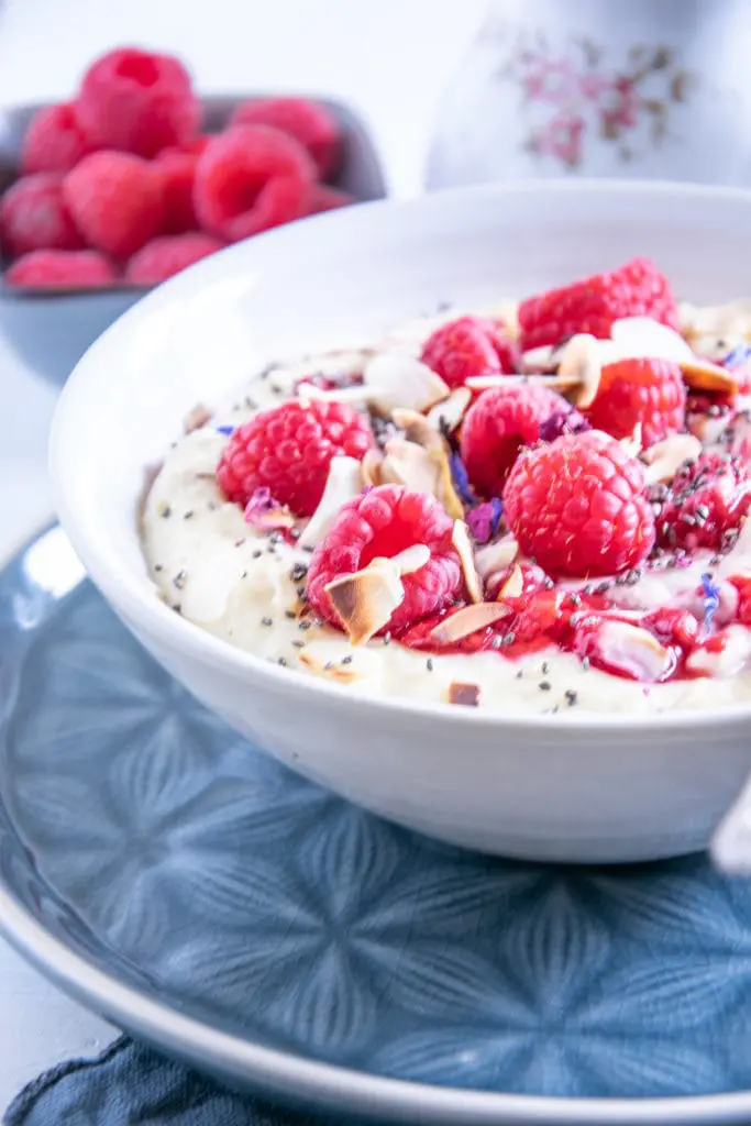 Vanillepudding-Porridge mit warmen Himbeeren in einer weißen Schüsse auf einem blauen Teller im 45-Grad-Winkel angeschnitten von rechts, mit Himbeeren, Mandelblättchen, Chiasamen und Blüten als Topping