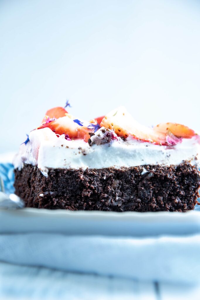 Ein Stück Brownie Style Erdbeerkuchen mit Topfen-Joghurtcreme auf einem dunkelblauen Teller mit Gabel auf Augenhöhe