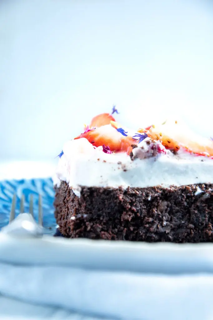 Ein Stück Brownie Style Erdbeerkuchen mit Topfen-Joghurtcreme auf einem dunkelblauen Teller mit Gabel auf Augenhöhe