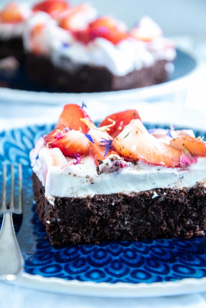 Ein Stück Brownie Style Erdbeerkuchen mit Topfen-Joghurtcreme auf einem dunkelblauen Teller mit Gabel im 45-Grad-Winkel