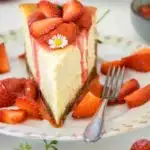 Ein Stück Cheesecake mit Erdbeerkompott auf einem altmodischen Kuchenteller mit Rosenmuster und Goldrand. Nahaufnahme im 45-Grad-Winkel mit Fokus auf das Kompott und das Gänseblümchen auf der Oberfläche des Tortenstücks. Erdbeersauce fließt an den Seitenrändern der cremigen Cheesecakemasse auf den Teller.