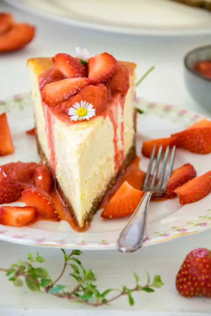 Ein Stück Cheesecake mit Erdbeerkompott auf einem altmodischen Kuchenteller mit Rosenmuster und Goldrand. Nahaufnahme im 45-Grad-Winkel mit Fokus auf das Kompott und das Gänseblümchen auf der Oberfläche des Tortenstücks. Erdbeersauce fließt an den Seitenrändern der cremigen Cheesecakemasse auf den Teller.