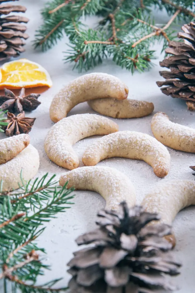 Gesündere Vanillekipferl auf weißem Untergrund dekoriert mit Tannenzweigen, Tannenzapfen, Anissternen und getrockneten Orangenscheiben, bestreut mit Puder-Erythrit. Aufnahme im 45-Grad-Winkel.