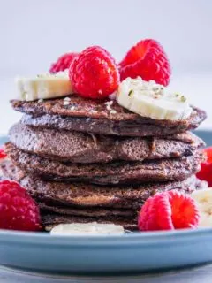 Ein Stapel Double Chocolate Protein-Pancakes garniert mit Bananenscheiben und Himbeeren. Fotografiert auf Augenhöhe.