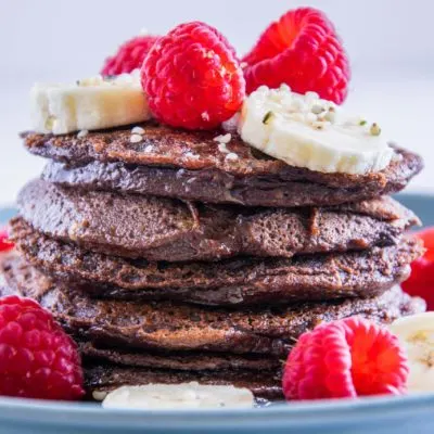 Ein Stapel Double Chocolate Protein-Pancakes garniert mit Bananenscheiben und Himbeeren. Fotografiert auf Augenhöhe.