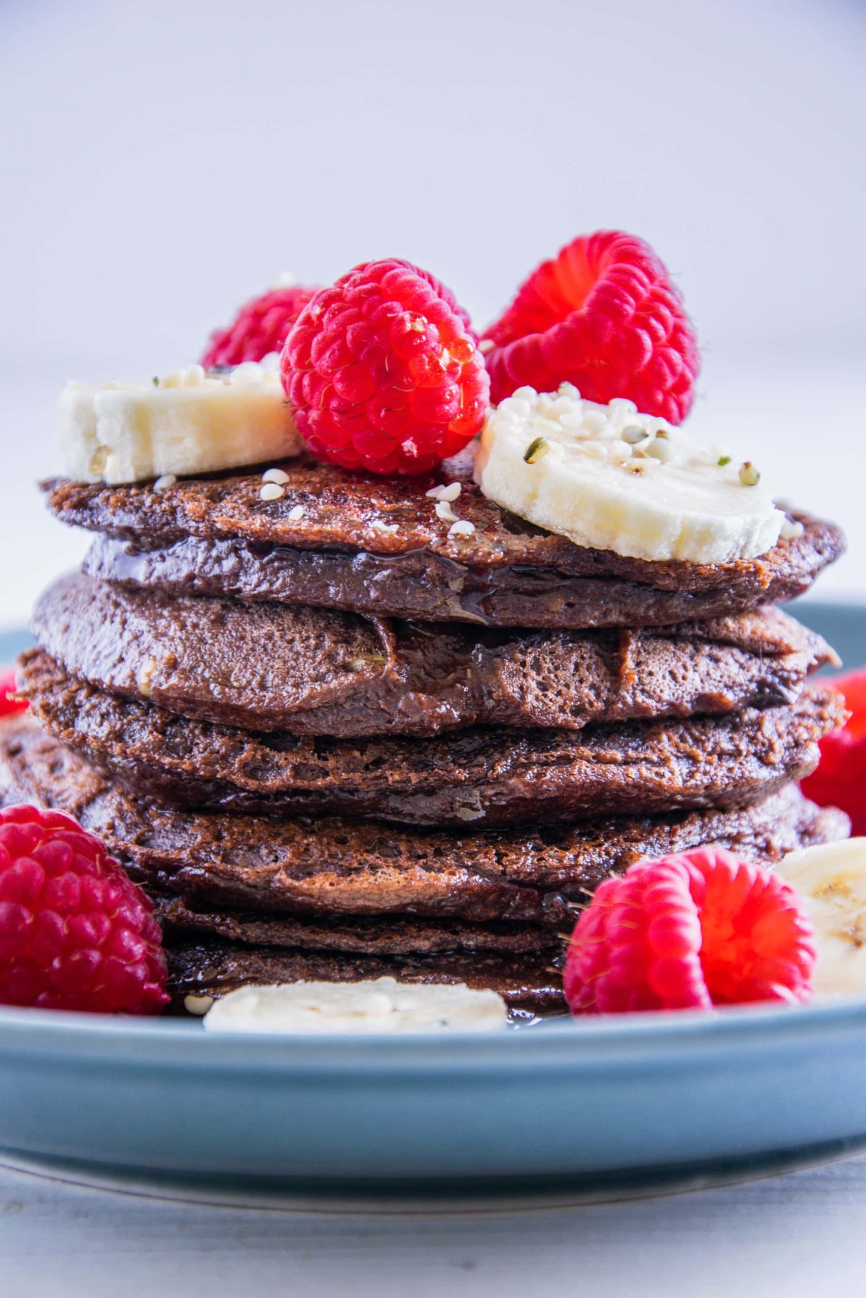 Ein Stapel Double Chocolate Protein-Pancakes garniert mit Bananenscheiben und Himbeeren. Fotografiert auf Augenhöhe.
