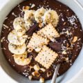 Cremiger Schokopudding-Porridge mit Schmelzflocken, Mini-Dinkelkeksen, Schokolade, gerösteten Macadamianüssen, Banane, Kokosflocken und Chiasamen