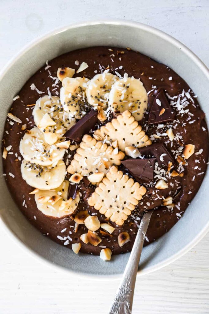 Cremiger Schokopudding-Porridge mit Schmelzflocken, Mini-Dinkelkeksen, Schokolade, gerösteten Macadamianüssen, Banane, Kokosflocken und Chiasamen