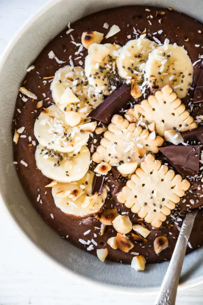 Cremiger Schokopudding-Porridge mit Schmelzflocken, Mini-Dinkelkeksen, Schokolade, gerösteten Macadamianüssen, Banane, Kokosflocken und Chiasamen