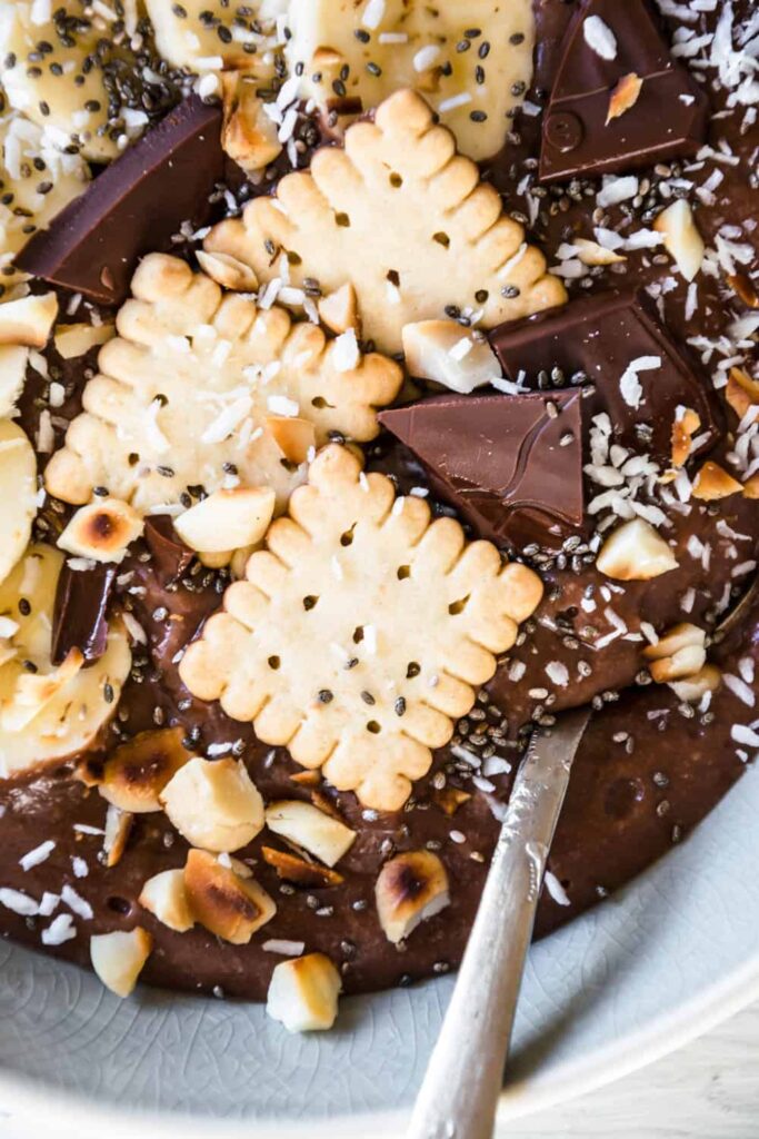 Cremiger Schokopudding-Porridge mit Schmelzflocken, Mini-Dinkelkeksen, Schokolade, gerösteten Macadamianüssen, Banane, Kokosflocken und Chiasamen