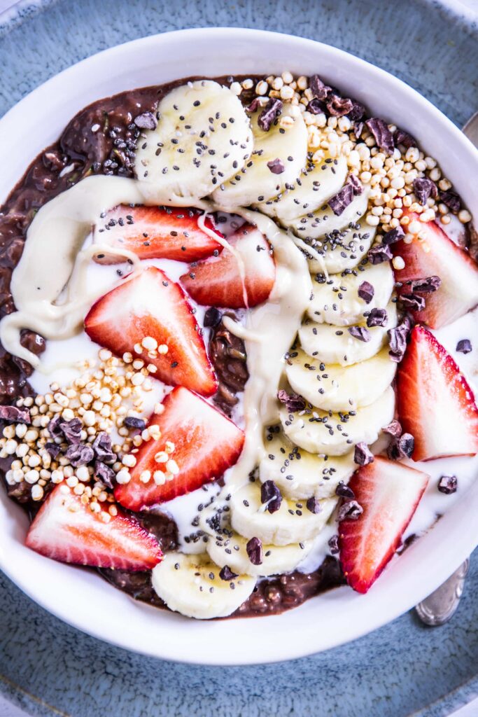 Banane-Schoko-Overnight-Zoats mit Erdbeeren, gepuffter Quinoa, Kakaonibs und in einem Bogen arrangierten Bananenscheiben als Topping in einer weißen Schüssel auf hellem Untergrund mit einem dunkeltürkisen Teller darunter.