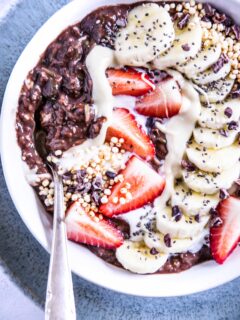 Banane-Schoko-Overnight-Zoats mit Erdbeeren, gepuffter Quinoa, Kakaonibs und in einem Bogen arrangierten Bananenscheiben als Topping in einer weißen Schüssel auf hellem Untergrund mit einem dunkeltürkisen Teller darunter.