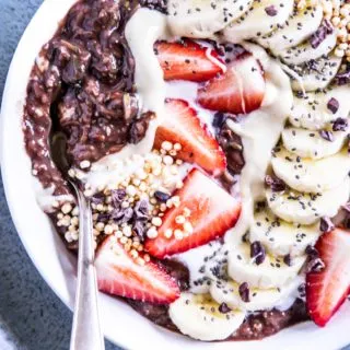 Banane-Schoko-Overnight-Zoats mit Erdbeeren, gepuffter Quinoa, Kakaonibs und in einem Bogen arrangierten Bananenscheiben als Topping in einer weißen Schüssel auf hellem Untergrund mit einem dunkeltürkisen Teller darunter.