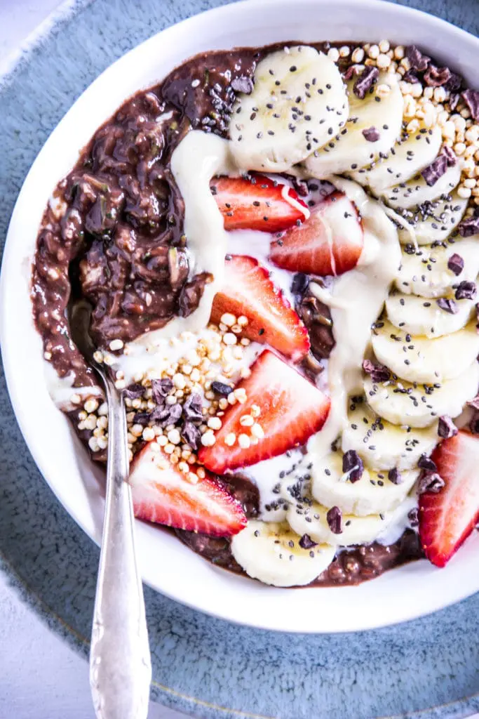 Banane-Schoko-Overnight-Zoats mit Erdbeeren, gepuffter Quinoa, Kakaonibs und in einem Bogen arrangierten Bananenscheiben als Topping in einer weißen Schüssel auf hellem Untergrund mit einem dunkeltürkisen Teller darunter.