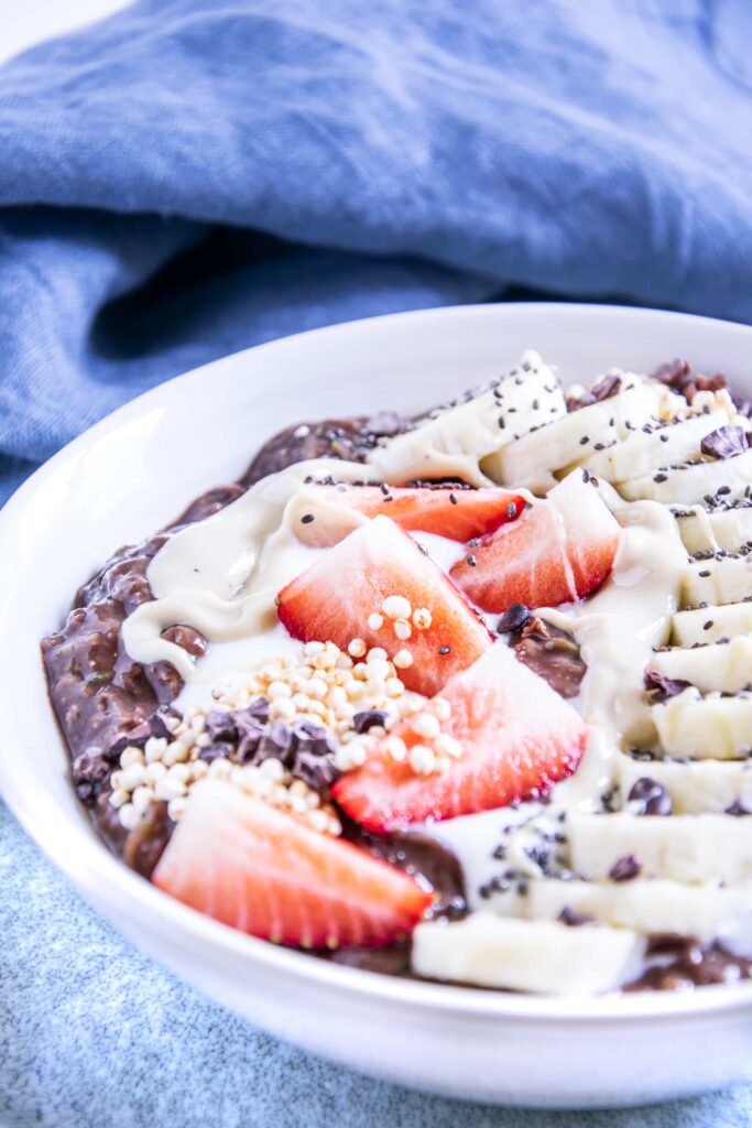 Banane-Schoko-Overnight-Zoats mit Erdbeeren, gepuffter Quinoa, Kakaonibs und in einem Bogen arrangierten Bananenscheiben als Topping in einer weißen Schüssel auf hellem Untergrund mit einem blauen Leinentuch