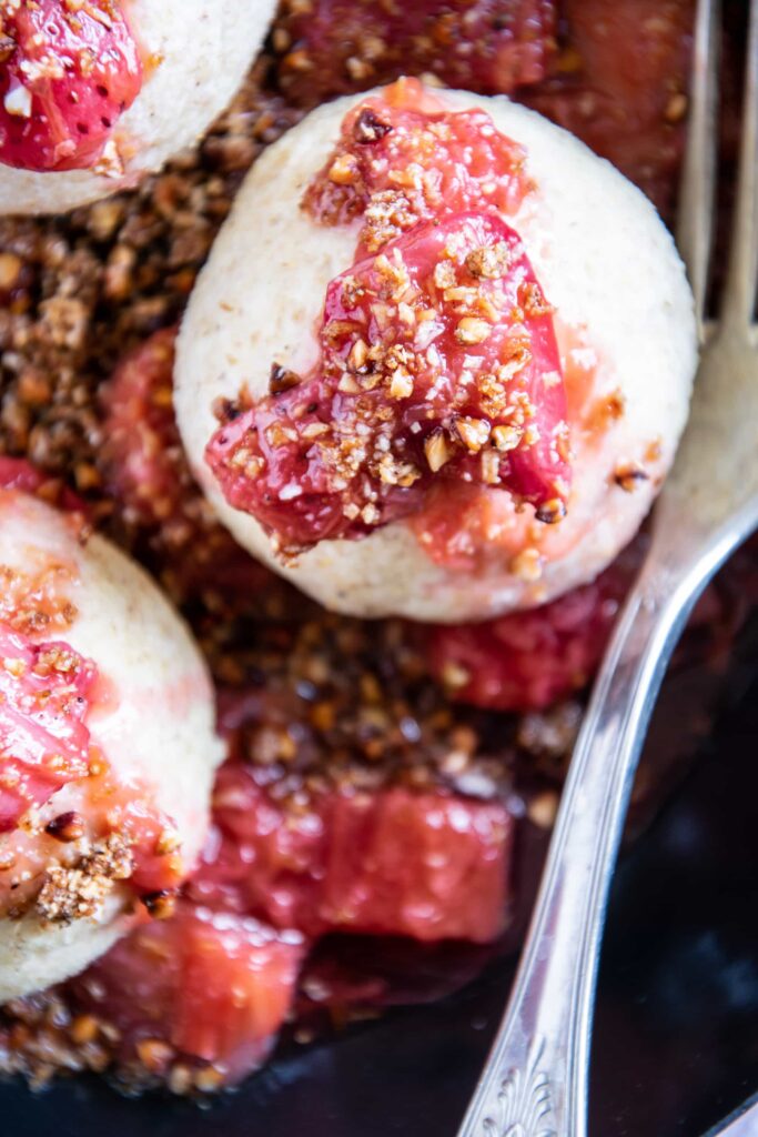 Ein Erdbeerknödel mit Buchweizen-Kokos-Crunch und Erdbeer-Rhabarberkompott plus Gabel in der Nahaufnahme.