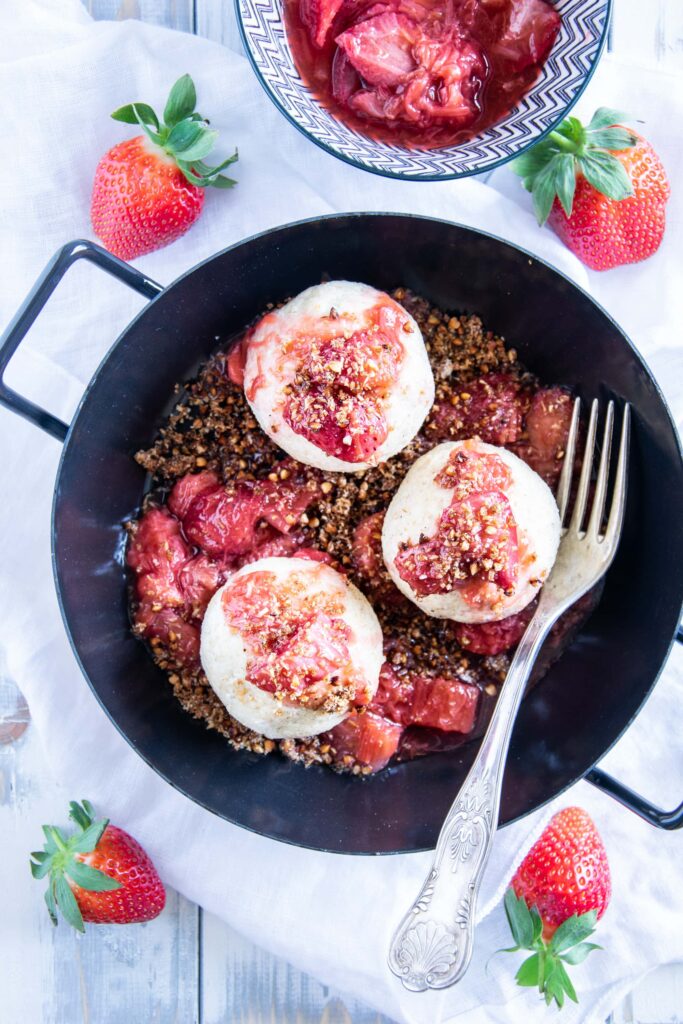 Gesunde Erdbeerknödel mit Buchweizen-Kokos-Crunch in einer schwarzen Servierpfanne mit zwei Henkeln. Heller Untergrund mit weißem Leinentuch, Gabel in der Pfanne und Erdbeeren als Deko