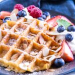 Knusprige Keto Zimt-Chaffles mit griechischem Sahnejoghurt, Beeren, gerösteten Mandelblättchen, Minze- und Melisseblättern auf einem schwarzen Teller mit einem dunkelblauen Leinentuch darunter. Aufnahme im 45°-Winkel.