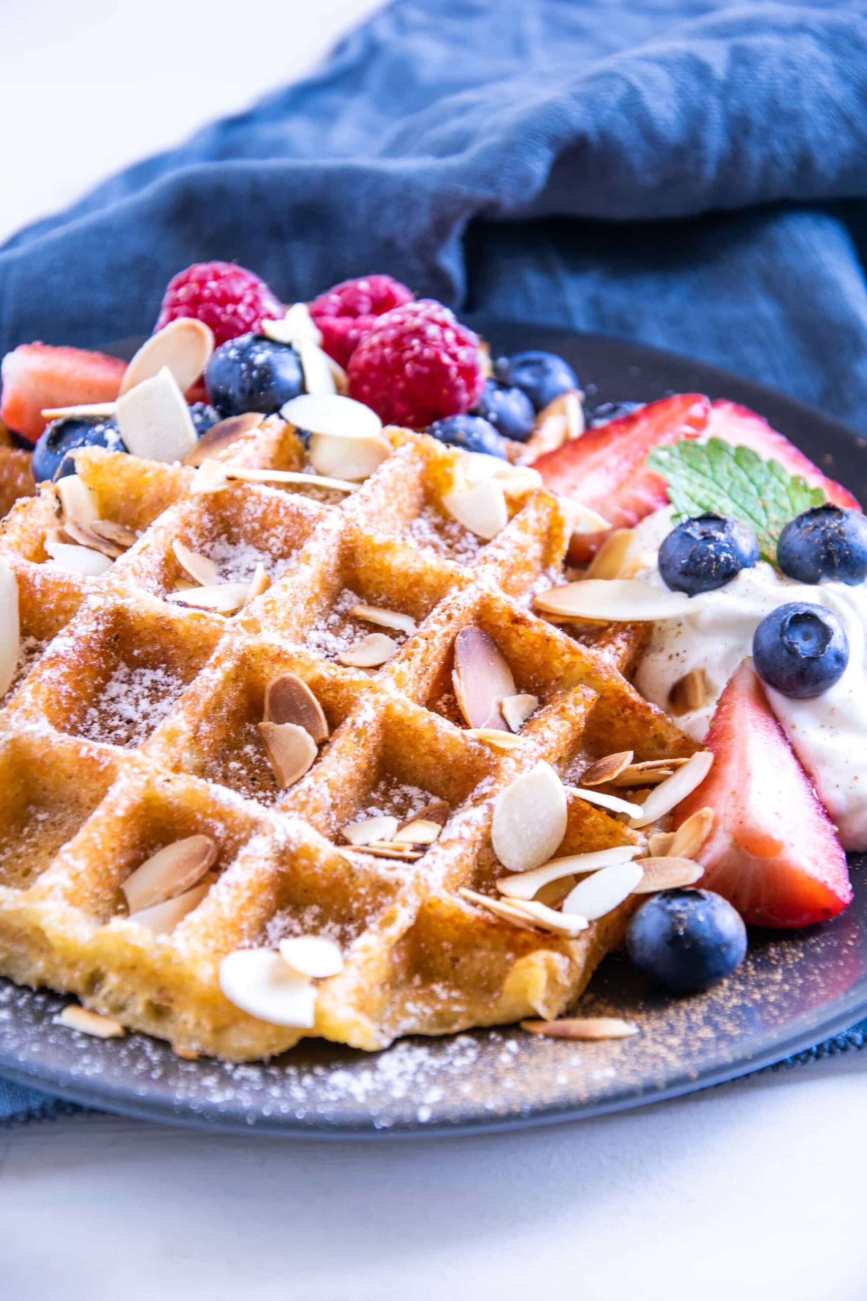 Knusprige Keto Zimt-Chaffles mit griechischem Sahnejoghurt, Beeren, gerösteten Mandelblättchen, Minze- und Melisseblättern auf einem schwarzen Teller mit einem dunkelblauen Leinentuch darunter. Aufnahme im 45°-Winkel.