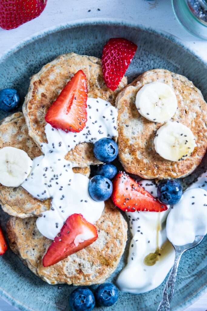 Hafer-Power-Pancakes in einem Bogen auf einem türkisen Teller angerichtet. Getoppt mit Joghurt, Beeren, Bananenscheiben und Chiasamen. Top View.