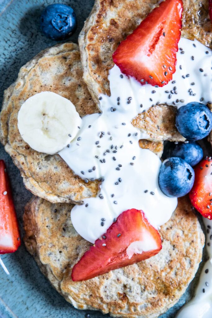 Hafer-Power-Pancakes in einem Bogen auf einem türkisen Teller angerichtet. Getoppt mit Joghurt, Beeren, Bananenscheiben und Chiasamen. Top View.