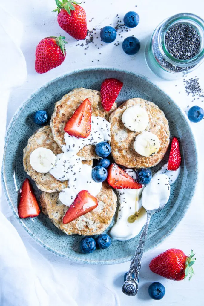 Hafer-Power-Pancakes in einem Bogen auf einem türkisen Teller angerichtet. Getoppt mit Joghurt, Beeren, Bananenscheiben und Chiasamen. Top View.