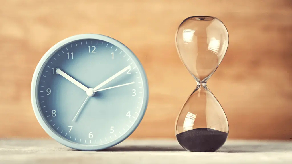Uhr und Sanduhr auf einem Tisch vor braunem Hintergrund.