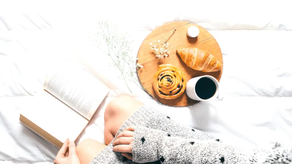 Flatlay mit einem aufgeschlagenen Buch, Knien und Händen der Person, die es liest, und einem Frühstücksteller mit süßem Gebäck und Kaffee.