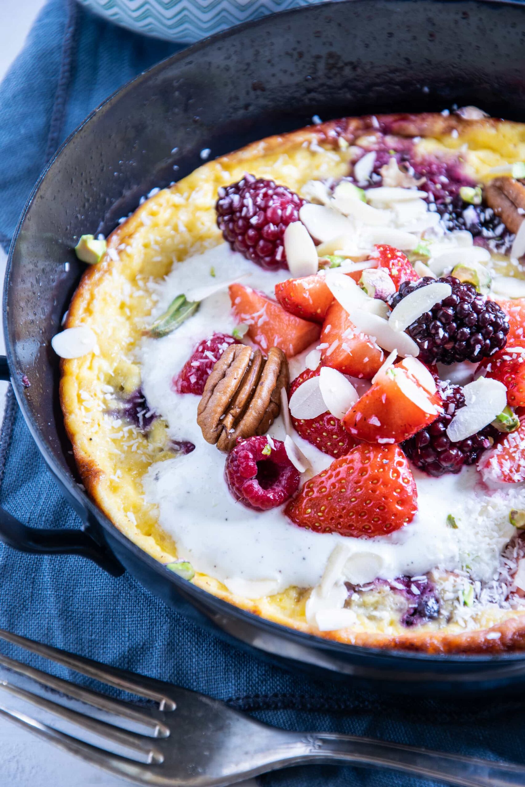 Süßes Keto-Omelette in einer schwarzen Gusseisen-Servierpfanne getoppt mit Mascarpone-Joghurtcreme, Beeren und Nüssen. Blaues Leinentuch und Top View.