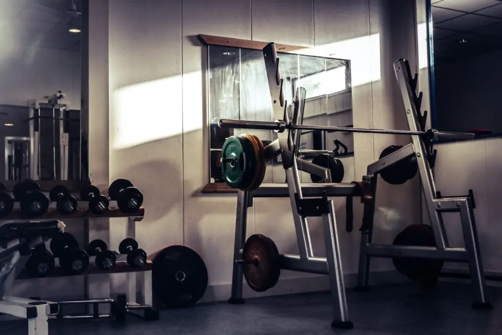 Squat Rack mit Langhanteln und Hantelscheiben. Spiegel im Hintergrund. Kurzhanteln auf einer Ablage daneben.
