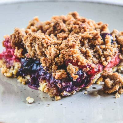 Ein Stück gesunder, zuckerfreier Beeren-Streuselkuchen auf einem grauen Teller. Ansicht im 45-Grad-Winkel. Sehr knusprige Textur.