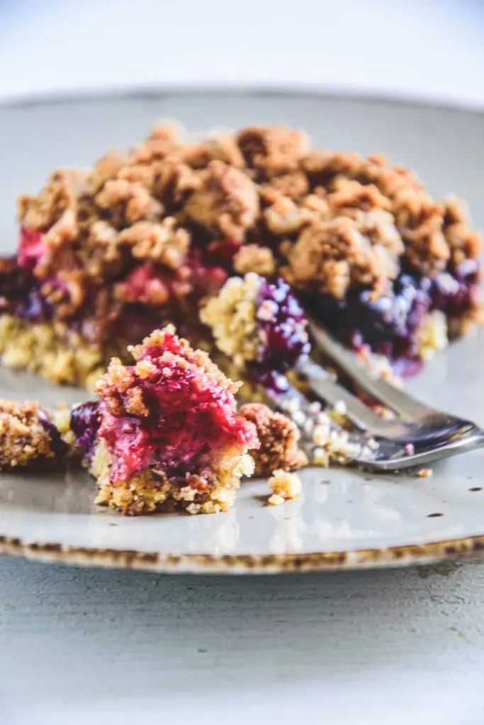 Ein Stück gesunder Beeren-Streuselkuchen mit Dinkelmehl und Mandeln, ohne Zucker. Ansicht im 45-Grad-Winkel. Eine Gabel hat ein Stück vom Kuchen abgestochen.
