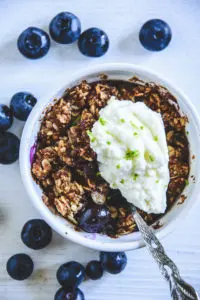 Blueberry Crumble mit Limetten-Quark in einer kleinen, weißen Portionsform mit Heidelbeeren rundherum. Top View.