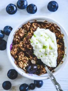 Blueberry Crumble mit Limetten-Quark in einer kleinen, weißen Portionsform mit Heidelbeeren rundherum. Top View.