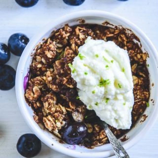 Blueberry Crumble mit Limetten-Quark in einer kleinen, weißen Portionsform mit Heidelbeeren rundherum. Top View.