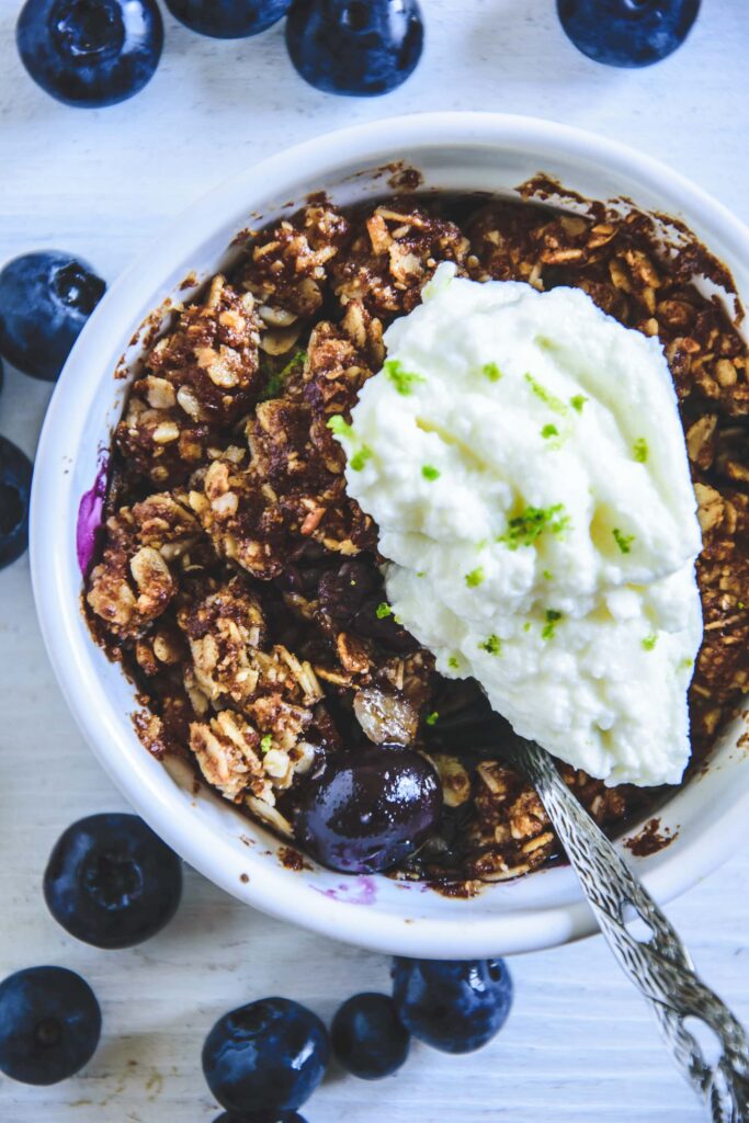 Blueberry Crumble mit Limetten-Quark in einer kleinen, weißen Portionsform mit Heidelbeeren rundherum. Top View.