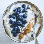 Heidelbeer-Kokos-Porridge in einer Schüssel getoppt mit Heidelbeeren, Mandelmus und Kokoschips. Top View. Löffel im Porridge.