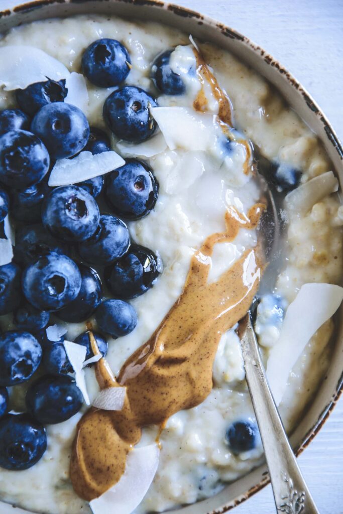 Heidelbeer-Kokos-Porridge in einer Schüssel getoppt mit Heidelbeeren, Mandelmus und Kokoschips. Top View. Löffel im Porridge.