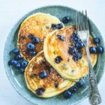 Heidelbeer-Topfen-Pancakes mit Gabel auf dunkeltürkisem Teller. Garniert mit Heidelbeeren. Weißer Untergrund. Top View.