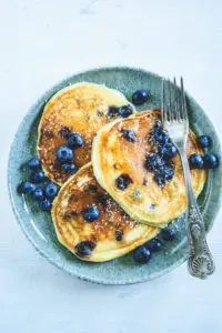 Heidelbeer-Topfen-Pancakes mit Gabel auf dunkeltürkisem Teller. Garniert mit Heidelbeeren. Weißer Untergrund. Top View.