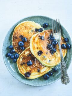 Heidelbeer-Topfen-Pancakes mit Gabel auf dunkeltürkisem Teller. Garniert mit Heidelbeeren. Weißer Untergrund. Top View.