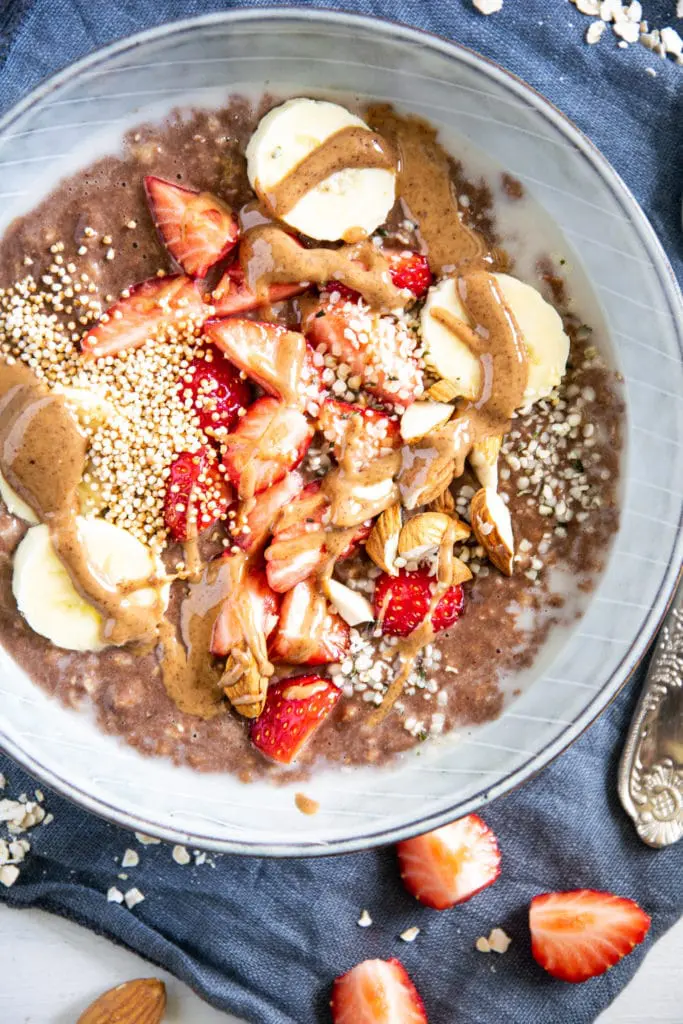 Cremiger Schoko-Mandel-Porridge getoppt mit Bananenscheiben, Erdbeeren, Mandeln und Mandelmus. In einer blaugrauen Schüssel mit Löffel darin, auf einem blauen Leinentuch. Top View.