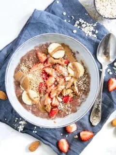 Cremiger Schoko-Mandel-Porridge getoppt mit Bananenscheiben, Erdbeeren, Mandeln und Mandelmus. In einer blaugrauen Schüssel mit Löffel darin, auf einem blauen Leinentuch. Top View.