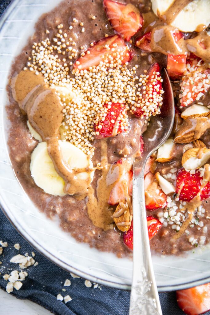 Nahansicht von Schoko-Mandel-Porridge getoppt mit Bananenscheiben, Erdbeeren, Mandeln und Mandelmus. In einer blaugrauen Schüssel mit Löffel darin. Top View.