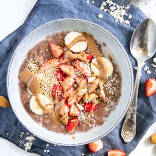 Cremiger Schoko-Mandel-Porridge getoppt mit Bananenscheiben, Erdbeeren, Mandeln und Mandelmus. In einer blaugrauen Schüssel mit Löffel darin, auf einem blauen Leinentuch. Top View.